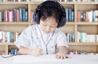 Un jeune enfant porte un casque et dessine sur une feuille de papier. Il se trouve devant une bibliothèque, avec des livres en arrière-plan.